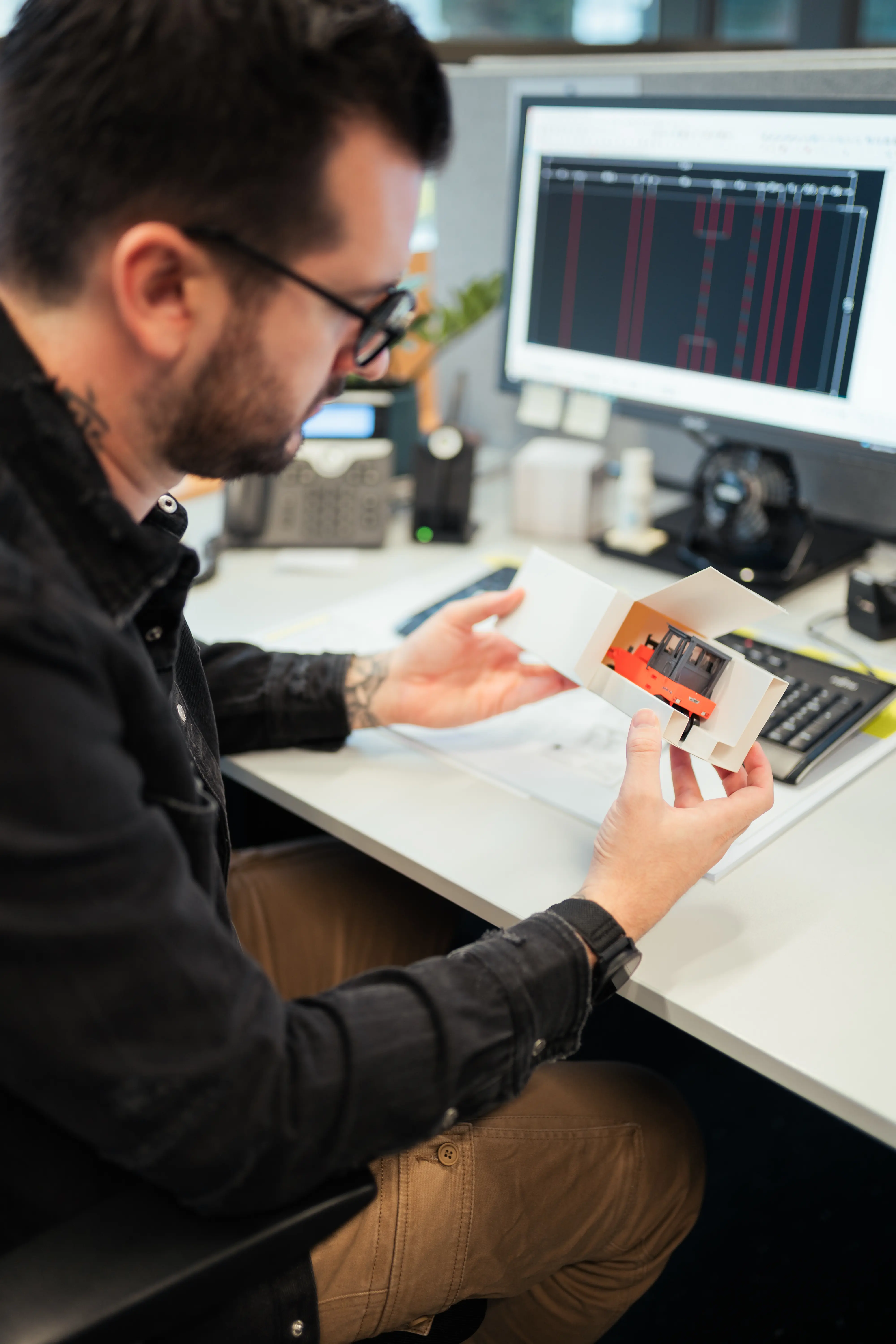 Verpackungstechniker prüft einen händisch gefertigten Prototypen einer Faltschachtel mit individuellem Inlay vor dem CAD-Bildschirm. Maßgeschneiderte Verpackungsentwicklung bei die-verpackungs-druckerei.